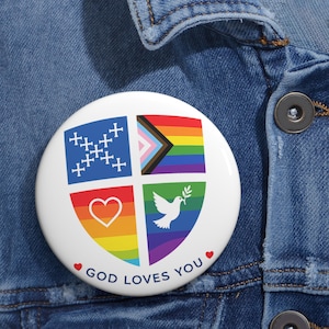 May include: A white button with a colorful design and the text "GOD LOVES YOU." The design includes a blue cross pattern, a rainbow flag, a heart, and a dove with an olive branch, all set against a rainbow background.