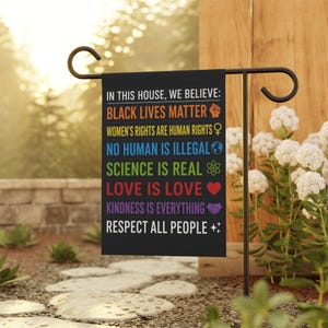 Könnte beinhalten: Eine schwarze Gartenflagge mit dem Text "IN THIS HOUSE, WE BELIEVE: BLACK LIVES MATTER, WOMEN'S RIGHTS ARE HUMAN RIGHTS, NO HUMAN IS ILLEGAL, SCIENCE IS REAL, LOVE IS LOVE, KINDNESS IS EVERYTHING, RESPECT ALL PEOPLE." Die Flagge wird im Freien gezeigt.