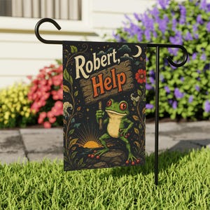 May include: A black garden flag with a green frog holding a sign saying "Robert, Help." The frog has red eyes and is on a rock. The background shows a sunset, flowers, and a crescent moon. The flag is displayed on a black metal stand.