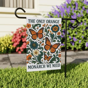 Puede incluir: Una bandera de jardín blanca con mariposas monarca naranjas, flores turquesas y hojas verdes. La bandera dice "THE ONLY ORANGE MONARCH WE NEED". Se muestra en un soporte de metal negro en un jardín.