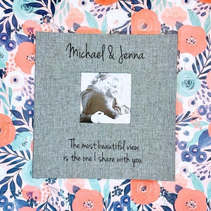 K&ouml;nnte beinhalten: Ein graues Fotoalbum mit einer schwarzen Aufschrift, die "Michael & Jenna" lautet. Das Album hat ein kleines Fenster mit einem Schwarzwei&szlig;foto eines Paares. Die Aufschrift unter dem Fenster lautet "Die sch&ouml;nste Aussicht, ist die, die ich mit dir teile".