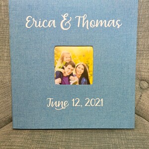 K&ouml;nnte beinhalten: Ein blaues Fotoalbum mit einem wei&szlig;en Fotorahmen und dem Text "Erica & Thomas" und "June 12, 2021". Der Fotorahmen enth&auml;lt ein Foto einer dreik&ouml;pfigen Familie.