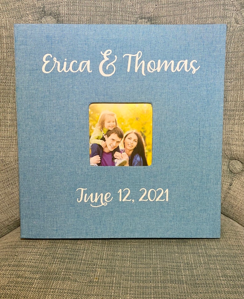 K&ouml;nnte beinhalten: Ein blaues Fotoalbum mit einem wei&szlig;en Fotorahmen und dem Text "Erica & Thomas" und "June 12, 2021". Der Fotorahmen enth&auml;lt ein Foto einer dreik&ouml;pfigen Familie.