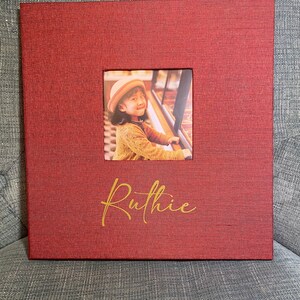 May include: A red photo album with a window showing a young girl wearing a red sweater and a white hat. The album has the name "Ruthie" written in gold lettering.