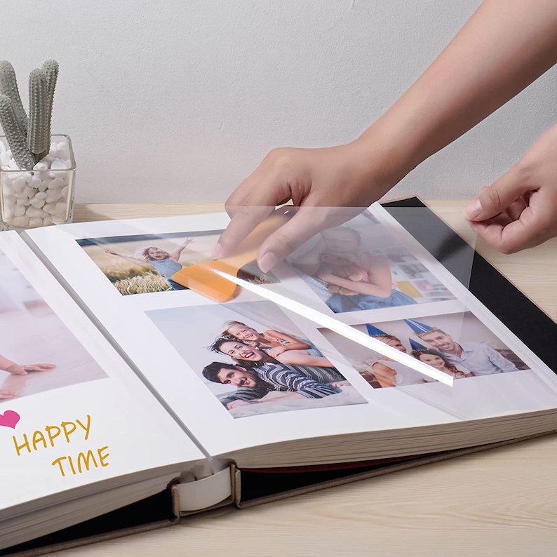 K&ouml;nnte beinhalten: Eine Person bringt eine transparente Plastikfolie auf ein Fotoalbum an. Das Album ist auf einer Seite mit mehreren Fotos einer Familie ge&ouml;ffnet. Auf der Seite steht auch der Text "Happy Time".