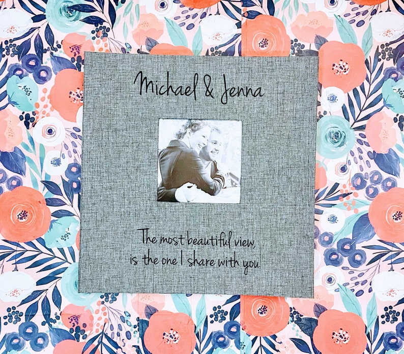 K&ouml;nnte beinhalten: Ein graues Fotoalbum mit einer schwarzen Aufschrift, die "Michael & Jenna" lautet. Das Album hat ein kleines Fenster mit einem Schwarzwei&szlig;foto eines Paares. Die Aufschrift unter dem Fenster lautet "Die sch&ouml;nste Aussicht, ist die, die ich mit dir teile".