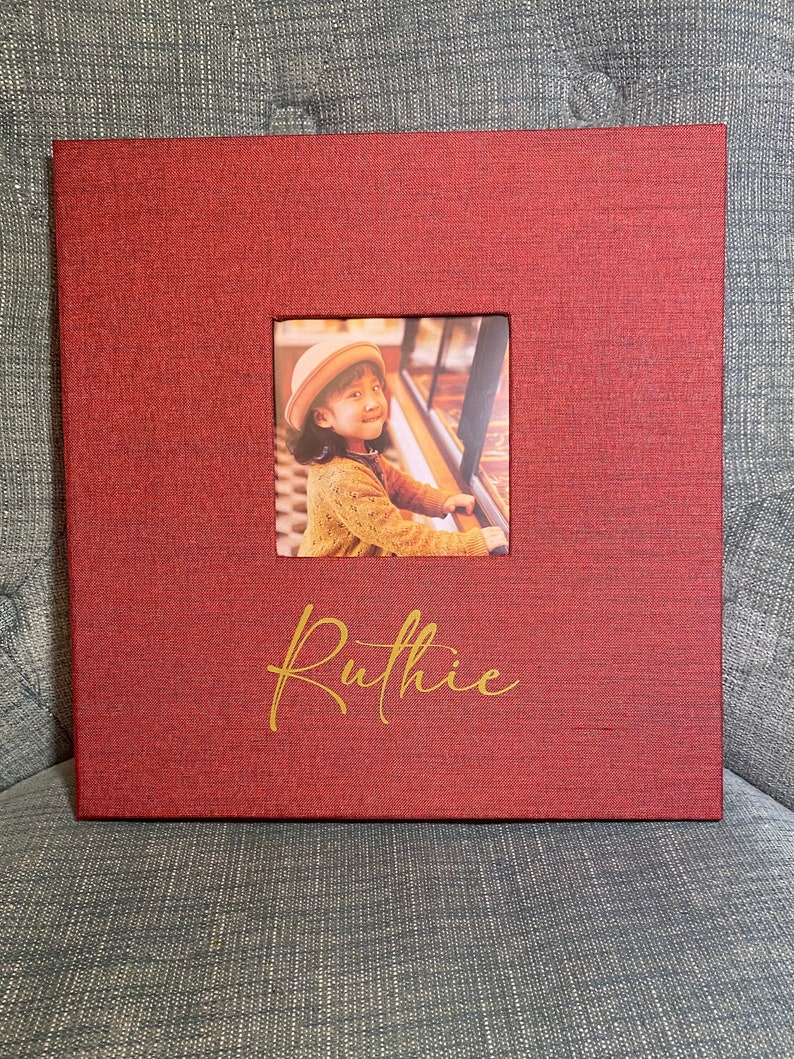 K&ouml;nnte beinhalten: Ein rotes Fotoalbum mit einem Fenster, das ein junges M&auml;dchen zeigt, das einen roten Pullover und eine wei&szlig;e M&uuml;tze tr&auml;gt. Das Album tr&auml;gt den Namen "Ruthie" in goldenen Lettern.