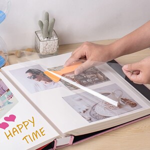 May include: A person is using a clear plastic sheet to protect photos in a pink photo album. The album has the words "Happy Time" written on the front.