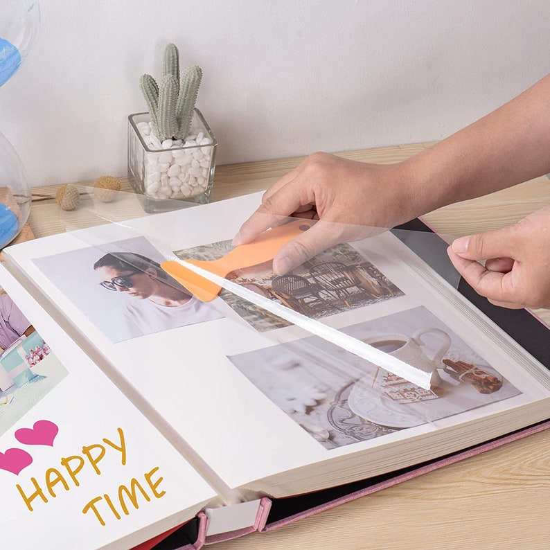 May include: A person is using a clear plastic sheet to protect photos in a pink photo album. The album has the words "Happy Time" written on the front.
