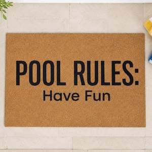 May include: A rectangular doormat with the text "POOL RULES: Have Fun" in black letters. The mat is brown and sits on a light-colored tile surface. Yellow and blue striped towels are visible.