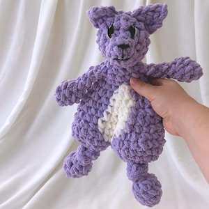 May include: A plush, hand-crocheted animal toy made from thick, soft lavender yarn. The toy features a white patch on its belly, pointed ears, and black plastic safety eyes. The texture is chunky and looped, creating a tactile, soft appearance.