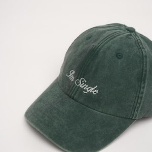 Könnte beinhalten: Eine dunkelwaldgrüne Baseballkappe mit verwaschenem Vintage-Finish. Auf der Vorderseite ist der Text "Im Single" in einer weißen Schreibschrift eingestickt. Die Kappe verfügt über einen gebogenen Schirm und einen Knopf in der Mitte oben.