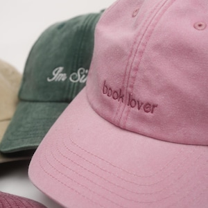 Könnte beinhalten: Eine hellrosa Baseballkappe mit dem in dunklerem Rosa gestickten Text book lover. Dahinter befindet sich eine dunkelgrüne Baseballkappe mit weißem Sticktext Im Si, und am unteren Rand ist ein Teil einer kastanienbraunen Kappe zu sehen.