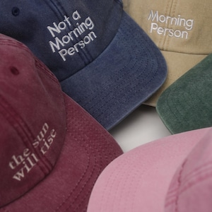 Könnte beinhalten: Eine Sammlung von fünf Baseballkappen in verschiedenen Farben, darunter Marineblau, Beige, Dunkelgrün, Kastanienbraun und Hellrosa. Die marineblaue Kappe trägt den weißen Sticktext Not a Morning Person, die beige Kappe Morning Person und die kastanienbraune the sun will rise.
