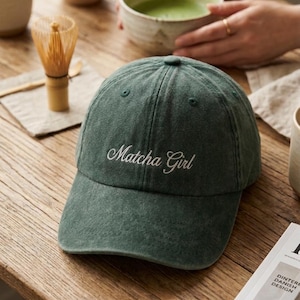 Op de afbeelding: Een donkergroene baseballpet met een verweerde stoftextuur en de tekst "Matcha Girl" geborduurd in wit cursief schrift op het voorpaneel. De pet heeft een gebogen klep, een knoop aan de bovenkant en gestikte oogjes voor ventilatie.