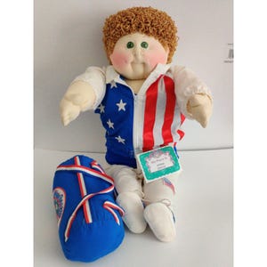 May include: A soft-sculpture doll with curly brown yarn hair and green eyes, wearing a white jacket with a blue star-patterned side and a red-and-white striped side. The doll wears white pants and shoes, and holds a blue duffel bag with a tag reading My Name Is Mark David.