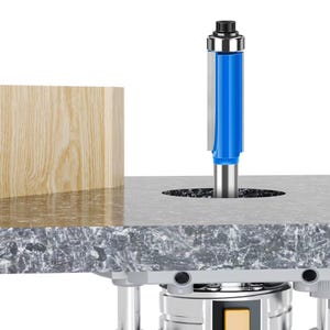 Puede incluir: Una fresa azul y plateada se inserta en una superficie gris, con un bloque de madera a la izquierda. La fresa es cilíndrica con un cuerpo azul y detalles plateados. El bloque de madera es marrón claro con vetas visibles.