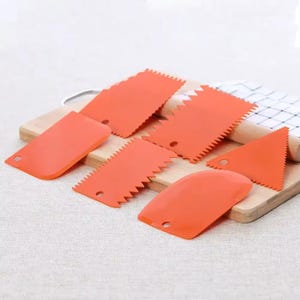 May include: An assortment of orange plastic cake decorating scrapers in various shapes, including straight edges, zigzags, and triangles. The scrapers are arranged on a wooden cutting board, ideal for smoothing frosting on cakes.