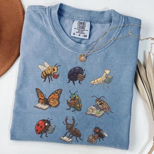 May include: A blue short-sleeved t-shirt featuring a graphic print of nine different insects, each reading a book. The insects include a bee, beetle, praying mantis, butterfly, ladybug, and others. The shirt has a Comfort Colors brand label.
