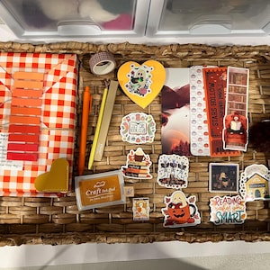 May include: A woven wicker tray containing a gift wrapped in orange and white gingham paper, a yellow heart-shaped notepad, an orange pen, a yellow pencil, a craft ink pad, and various stickers featuring ghosts, books, and autumn themes.