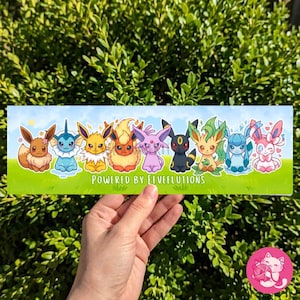 May include: A rectangular sticker featuring cartoon characters, including Eevee and its evolutions, on a green field under a blue sky. The text "POWERED BY EEVEEUTIONS" is at the bottom. The sticker is held by a hand.