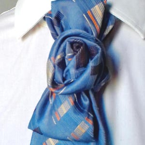 May include: A blue tie with a plaid pattern in orange, white, and black is knotted decoratively. The tie is worn with a white collared shirt. The tie's knot is approximately 10 cm in diameter.