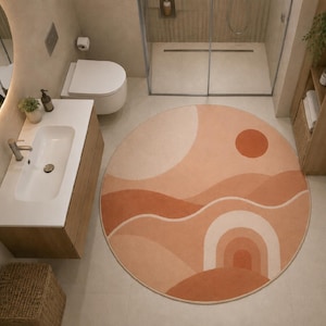May include: A round area rug featuring a desert-inspired landscape pattern in shades of terracotta, peach, and cream. The design includes abstract rolling hills, a solid circular sun, and a series of concentric arches on a light-colored floor.