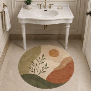 Op de afbeelding: Een ronde vloerkleed met een abstract landschapsontwerp. Het vloerkleed heeft een beige achtergrond met groene, oranje en bruine vormen, waaronder een zon en een plant. Het vloerkleed ligt op een betegelde vloer voor een witte wastafel.