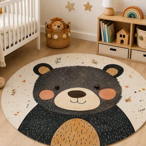 May include: A round area rug featuring a large, centered illustration of a black bear face with a tan muzzle, round pink cheeks, and small ears. The background is cream-colored with scattered small orange and brown leaf and dot patterns.