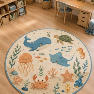 Könnte beinhalten: Runder Teppich mit Meeresmotiven. Er zeigt Illustrationen von Wal, Schildkröte, Qualle, Rochen, Krebs, Seestern und Seepferdchen. Der Teppich hat einen hellbeigen Hintergrund und einen blauen Rand. Durchmesser ca. 150 cm.