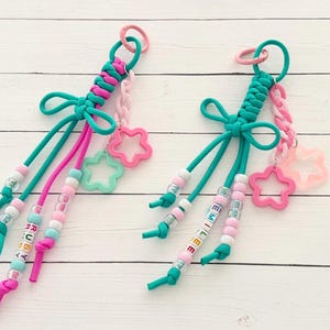 May include: Two handmade keychains with teal, pink, and white accents. Each keychain features a braided top, dangling cords, beads, and star-shaped charms. One keychain spells "RUBY" and the other spells "EMILE" with letter beads.