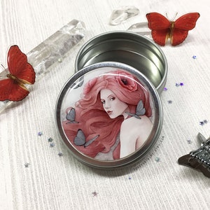 May include: A silver metal tin with a round lid. The lid has a colorful image of a woman with pink hair and a poppy flower.  She is surrounded by butterflies. The text "FOREVERFAIRY ETSY.COM" is printed on the lid.