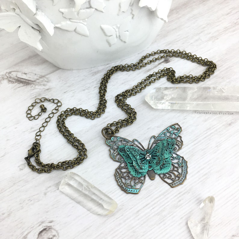 Puede incluir: Un collar de color bronce con un colgante de mariposa detallado con detalles en turquesa y bronce. La mariposa tiene un dise&ntilde;o de filigrana y un peque&ntilde;o cristal. El collar se exhibe sobre una superficie de madera clara con cristales y un objeto decorativo blanco.