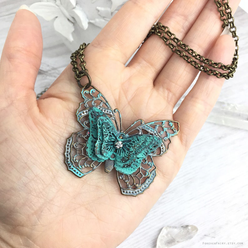 Puede incluir: Collar con colgante de mariposa detallado, con acabado turquesa y bronce. La mariposa tiene intrincadas alas caladas y un peque&ntilde;o cristal en el centro. La cadena del collar es de color bronce. El colgante se muestra en una mano.