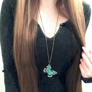 Puede incluir: Un collar largo de color oro antiguo con un colgante de mariposa turquesa. El colgante de mariposa tiene un dise&ntilde;o de filigrana y est&aacute; adornado con peque&ntilde;as piedras transparentes. El collar se lleva sobre un top negro acanalado.