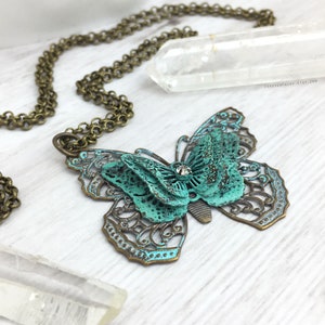 Puede incluir: Un collar con colgante de mariposa en tono bronce con p&aacute;tina turquesa. La mariposa tiene intrincadas alas de filigrana y un cristal central. La cadena del collar tambi&eacute;n es de color bronce. El fondo incluye un cristal transparente y una superficie blanca.