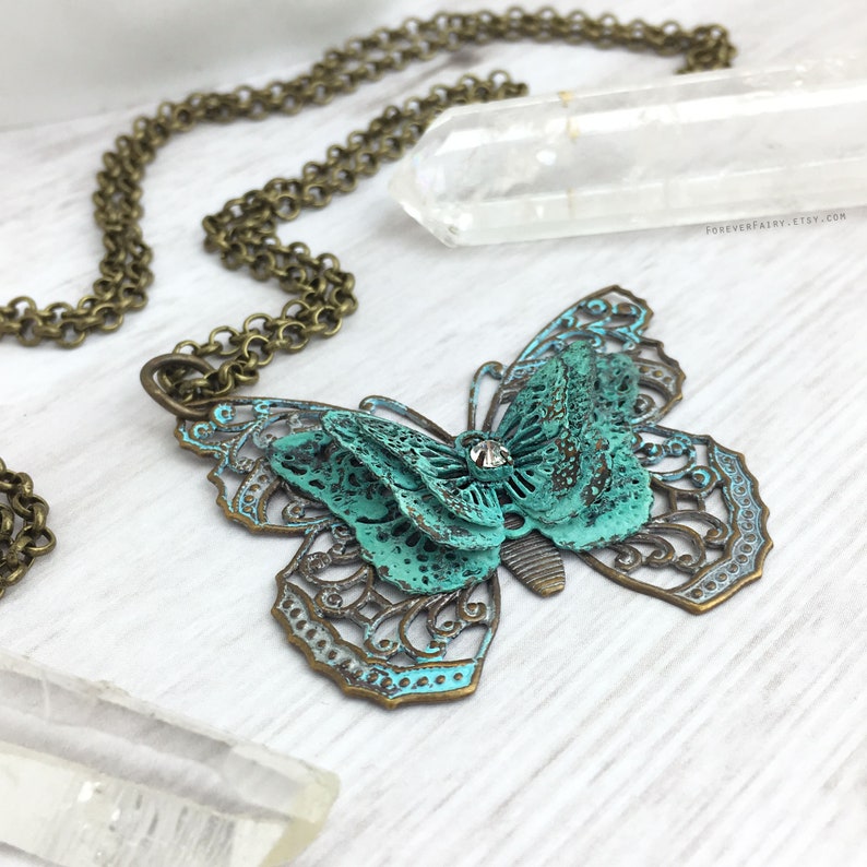 Puede incluir: Un collar con colgante de mariposa en tono bronce con p&aacute;tina turquesa. La mariposa tiene intrincadas alas de filigrana y un cristal central. La cadena del collar tambi&eacute;n es de color bronce. El fondo incluye un cristal transparente y una superficie blanca.