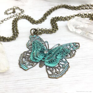 Puede incluir: Un collar con colgante de mariposa en tono bronce con centro turquesa y alas de filigrana. La mariposa tiene una peque&ntilde;a gema transparente en el centro. La cadena del collar tambi&eacute;n es de color bronce. El fondo es una superficie de madera clara.