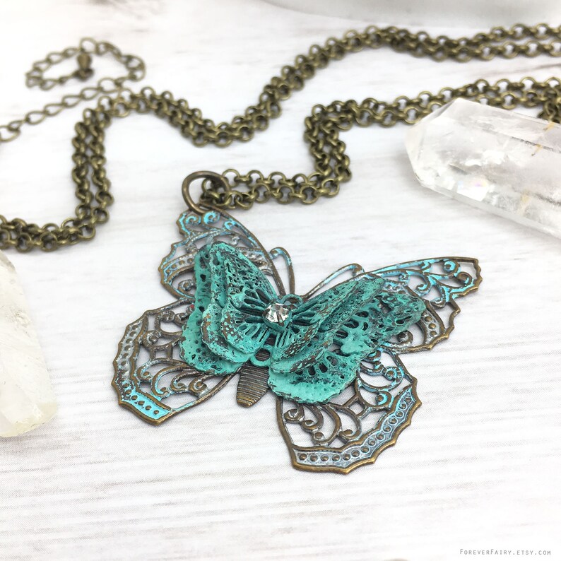 Puede incluir: Un collar con colgante de mariposa en tono bronce con centro turquesa y alas de filigrana. La mariposa tiene una peque&ntilde;a gema transparente en el centro. La cadena del collar tambi&eacute;n es de color bronce. El fondo es una superficie de madera clara.