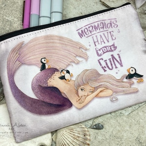 May include: A white zippered pouch featuring a purple mermaid with long flowing hair and a tail. The mermaid is surrounded by three small puffins. The text "Mermaids Have More Fun" is printed in purple on the pouch.