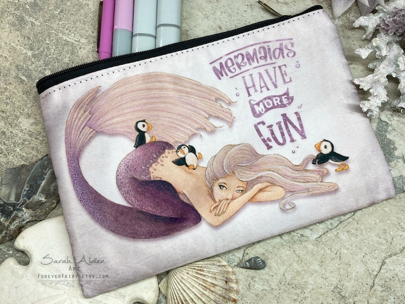 May include: A white zippered pouch featuring a purple mermaid with long flowing hair and a tail. The mermaid is surrounded by three small puffins. The text "Mermaids Have More Fun" is printed in purple on the pouch.