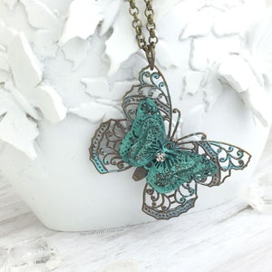 Puede incluir: Un collar con colgante de mariposa en color verde azulado y bronce. La mariposa tiene intrincadas alas de filigrana y un cuerpo texturizado con un peque&ntilde;o acento de cristal. La cadena del collar es de color bronce. El nombre de la tienda Etsy "foreverfairy.etsy.com" es visible.