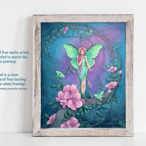 May include: A framed print of a watercolor painting featuring a fairy with green wings and a long green dress standing on a branch with pink flowers. The background is a dark blue with a crescent moon shape and green leaves. The print is surrounded by a white wooden frame.