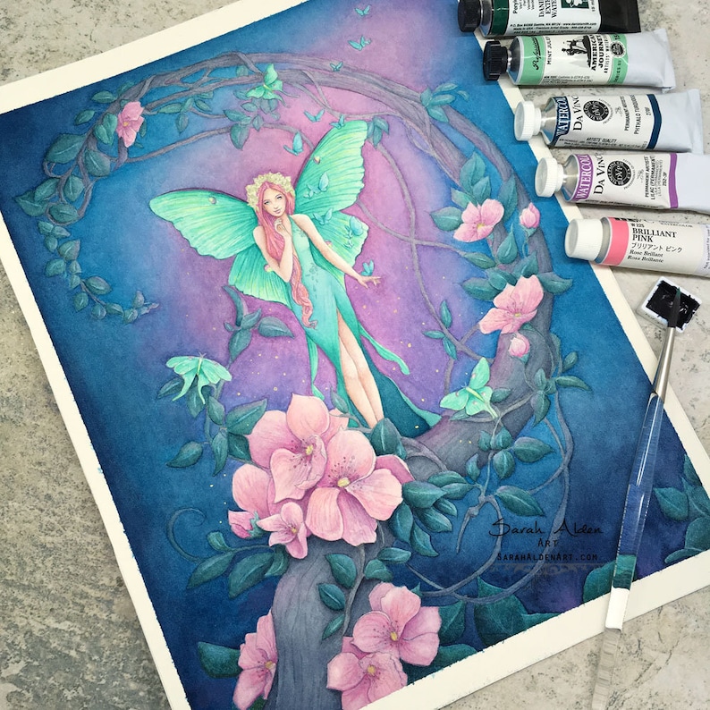 May include: Watercolor painting of a fairy with green wings and a pink dress standing on a tree branch with pink flowers. The background is a dark blue with purple and pink hues. The painting is surrounded by tubes of watercolor paint.