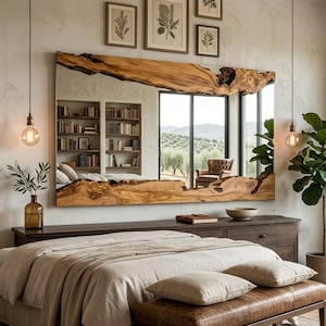 Puede incluir: Un gran espejo rectangular de pared con un marco de madera natural de borde vivo con bordes orgánicos e irregulares. El espejo refleja una habitación con una estantería, una ventana con vista a árboles y una silla. Se aprecian tonos marrones.