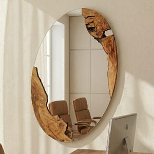 Puede incluir: Un espejo de pared ovalado con un marco de madera natural de borde vivo y veteado prominente. El borde de madera envuelve las secciones superior derecha e inferior izquierda de la superficie reflectante, creando un contorno orgánico asimétrico.