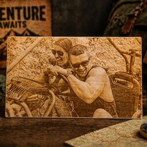 May include: A wooden photo with a laser-etched image of two people in sunglasses smiling. The photo is on a wooden surface with a lantern, a mug that says "Explore More", and a map in the background. The words "Adventure Awaits" are on a wooden sign.