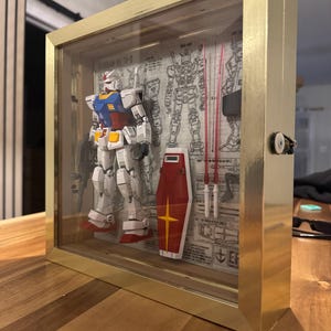 May include: A framed Gundam model in a gold-colored shadow box. The model is white, blue, red, and yellow, with a shield and weapons. The background features blueprints and technical drawings. The frame has a glass front.