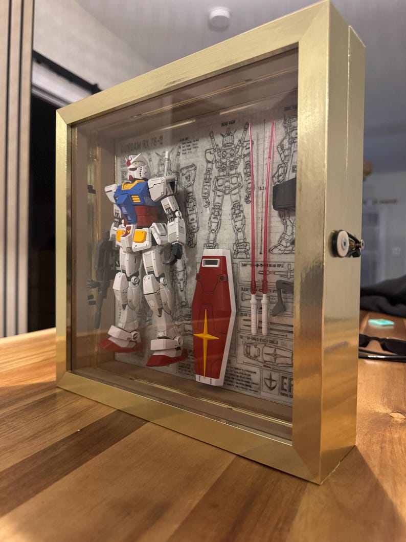 May include: A framed Gundam model in a gold-colored shadow box. The model is white, blue, red, and yellow, with a shield and weapons. The background features blueprints and technical drawings. The frame has a glass front.