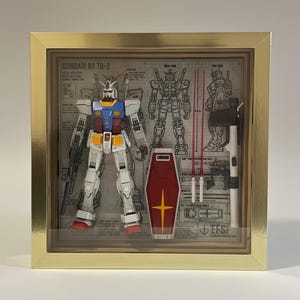 May include: A framed Gundam RX 78-2 model kit. The robot is white, blue, red, and yellow. Included are a red shield with a yellow cross, two red beam sabers, and a rifle. The background features blueprints.